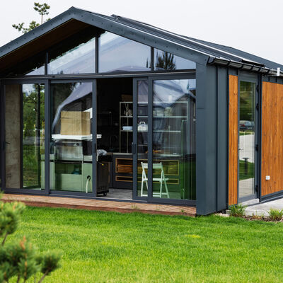 Modernes Tiny House mit Holzverkleidung, dunklem Stahlrahmen und gro&szlig;er Glasfront in einem gr&uuml;nen Garten